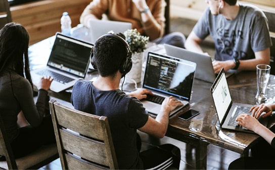 画像：日の入るオフィス、木目のテーブルに5人の人がノートパソコン、
イヤホンやヘッドフォンをしてソースを書いている様子。